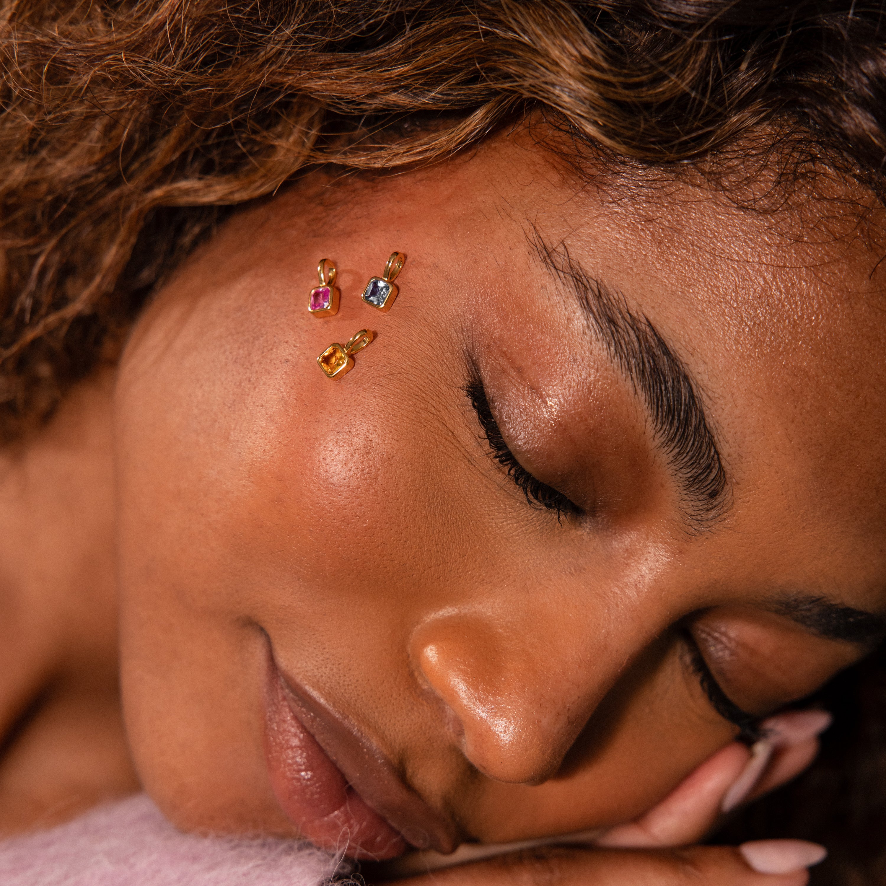Woman resting her face on her hand, eyes closed and serene, showcases the elegant Square Bezel Pink Tourmaline Charm as colorful gemstone jewelry placed gently on her cheek.