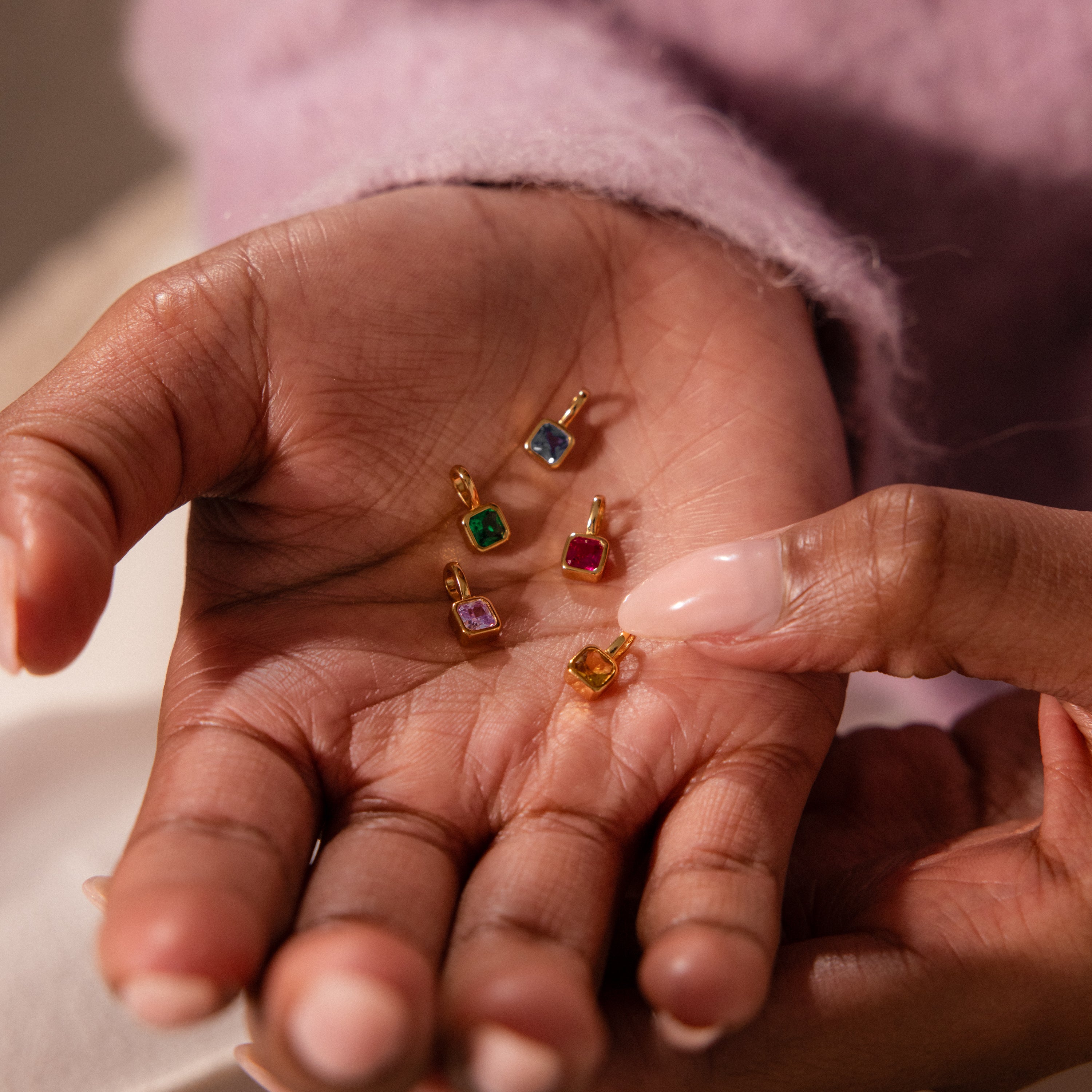 A hand holds four Square Bezel Birthstone Charms in gold frames, each featuring a vibrant gemstone, while another hand points at the colorful jewelry.
