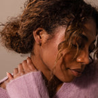 Woman with curly hair in a lavender sweater, eyes closed and hand on neck, wearing Amelia Duo Amethyst Huggies.