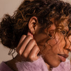 A woman with curly hair wears modern earrings—Amelia Duo Emerald Huggies—and a soft purple sweater, gently touching her ear.