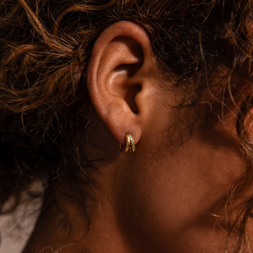 Close-up of a person’s ear with curly hair, wearing the Amelia Duo Blue Zircon Huggies—modern earrings featuring delicate gold hoops with sleek blue zircon accents.