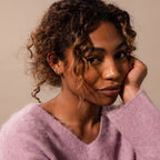 A woman with curly hair, in a soft purple sweater, smiles gently at the camera with her chin on her hand, showcasing elegant Amelia Duo Topaz Huggies.