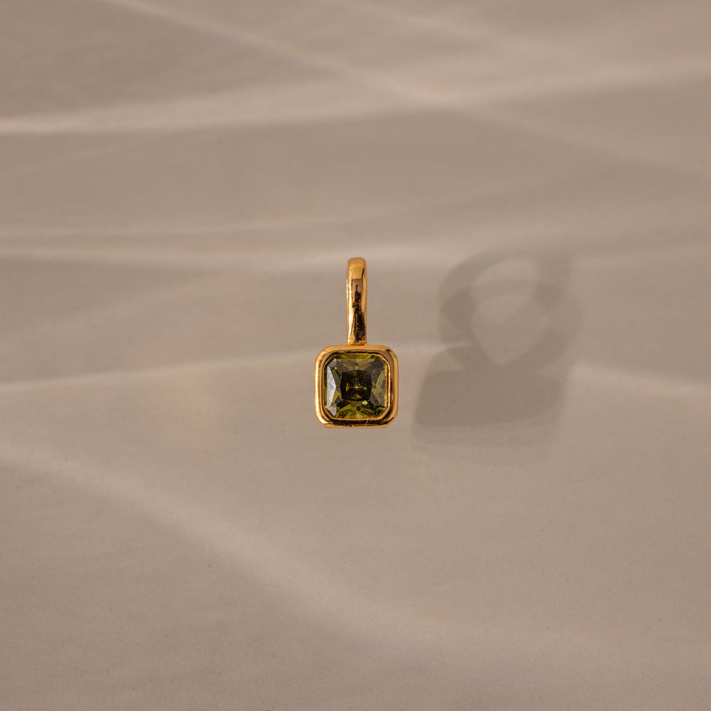 The Square Bezel Peridot Charm features a square green gemstone set in a gold frame, casting a shadow on a beige textured surface.