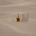 The Square Bezel Peridot Charm features a square green gemstone set in a gold frame, casting a shadow on a beige textured surface.