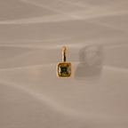 The Square Bezel Peridot Charm features a square green gemstone set in a gold frame, casting a shadow on a beige textured surface.