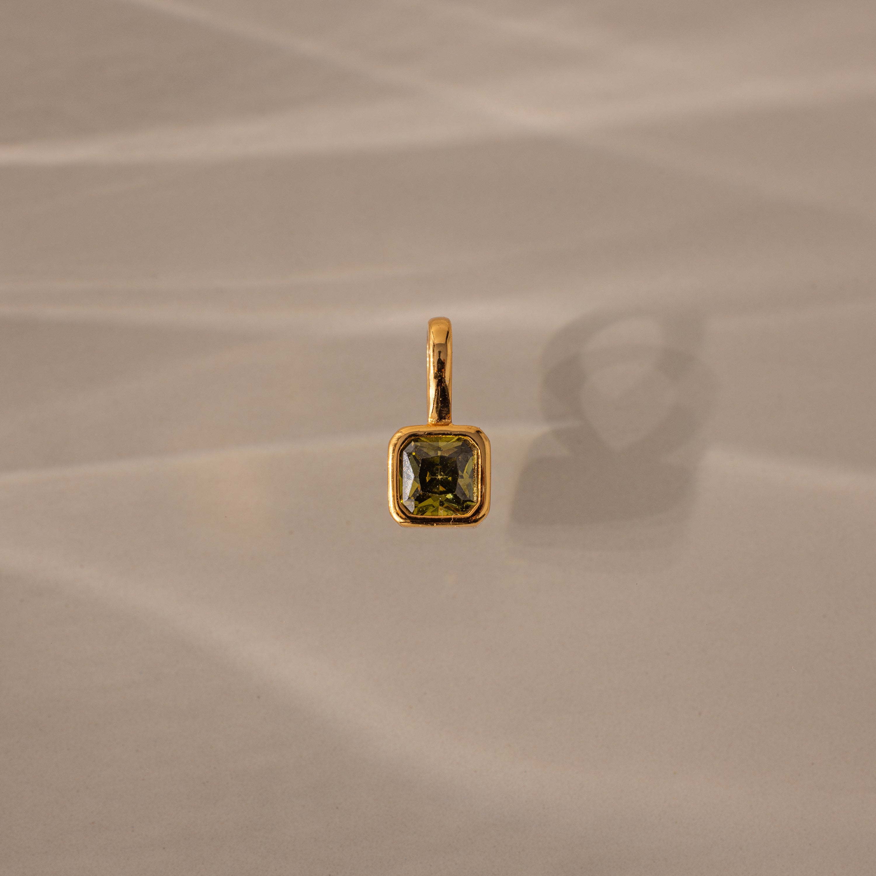 The Square Bezel Peridot Charm features a square green gemstone set in a gold frame, casting a shadow on a beige textured surface.