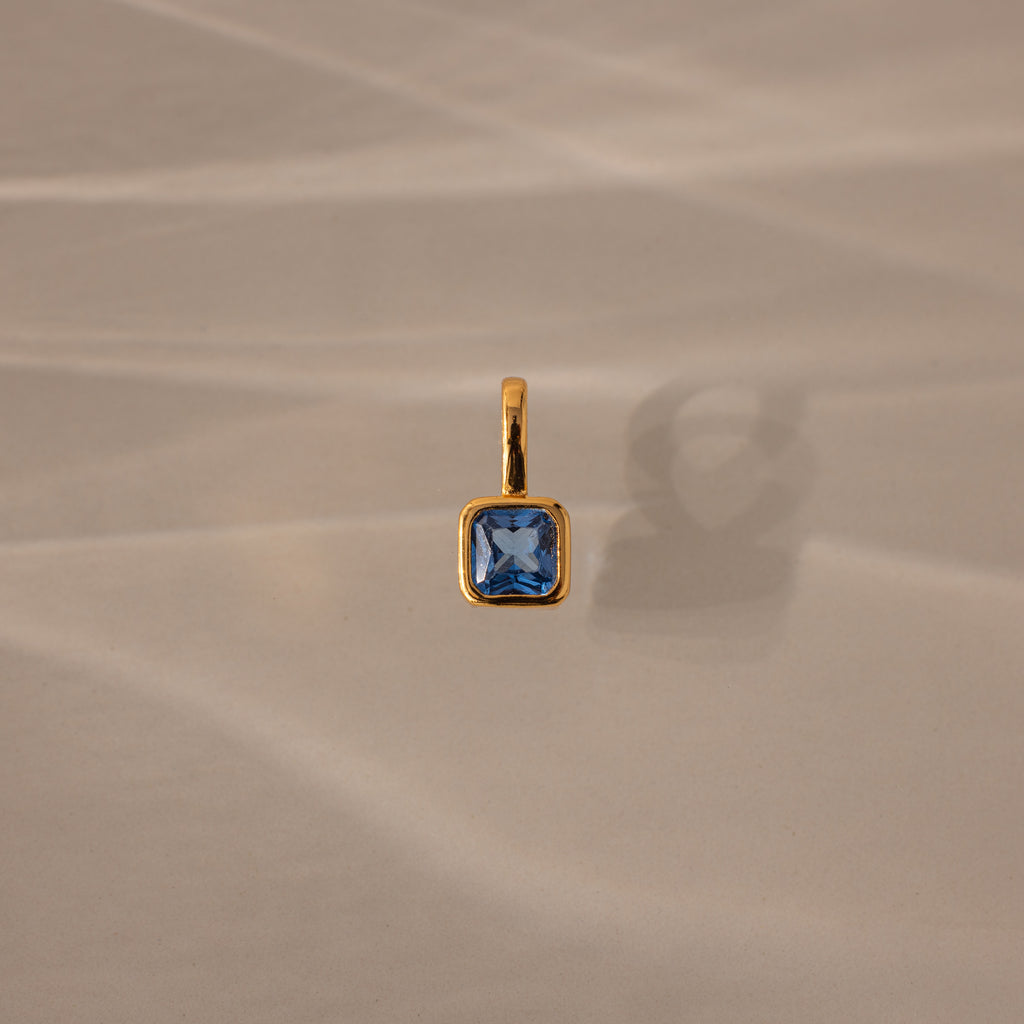 The Square Bezel Blue Zircon Charm features a square blue zircon set in a gold bezel, casting a soft shadow on beige textured fabric—a sophisticated piece of gemstone jewelry.