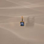 The Square Bezel Blue Zircon Charm features a square blue zircon set in a gold bezel, casting a soft shadow on beige textured fabric—a sophisticated piece of gemstone jewelry.
