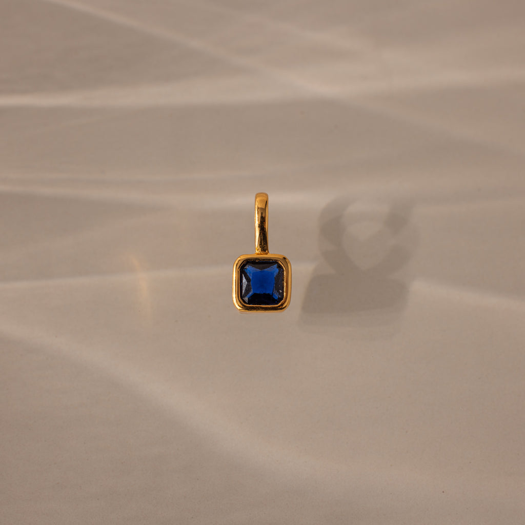 The Square Bezel Sapphire Charm, featuring a blue square gemstone set in a delicate gold frame, is ideal as a birthstone pendant and is shown resting on a beige surface with soft shadows.
