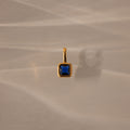 The Square Bezel Sapphire Charm, featuring a blue square gemstone set in a delicate gold frame, is ideal as a birthstone pendant and is shown resting on a beige surface with soft shadows.