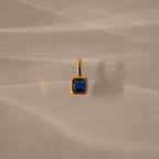 The Square Bezel Sapphire Charm, featuring a blue square gemstone set in a delicate gold frame, is ideal as a birthstone pendant and is shown resting on a beige surface with soft shadows.