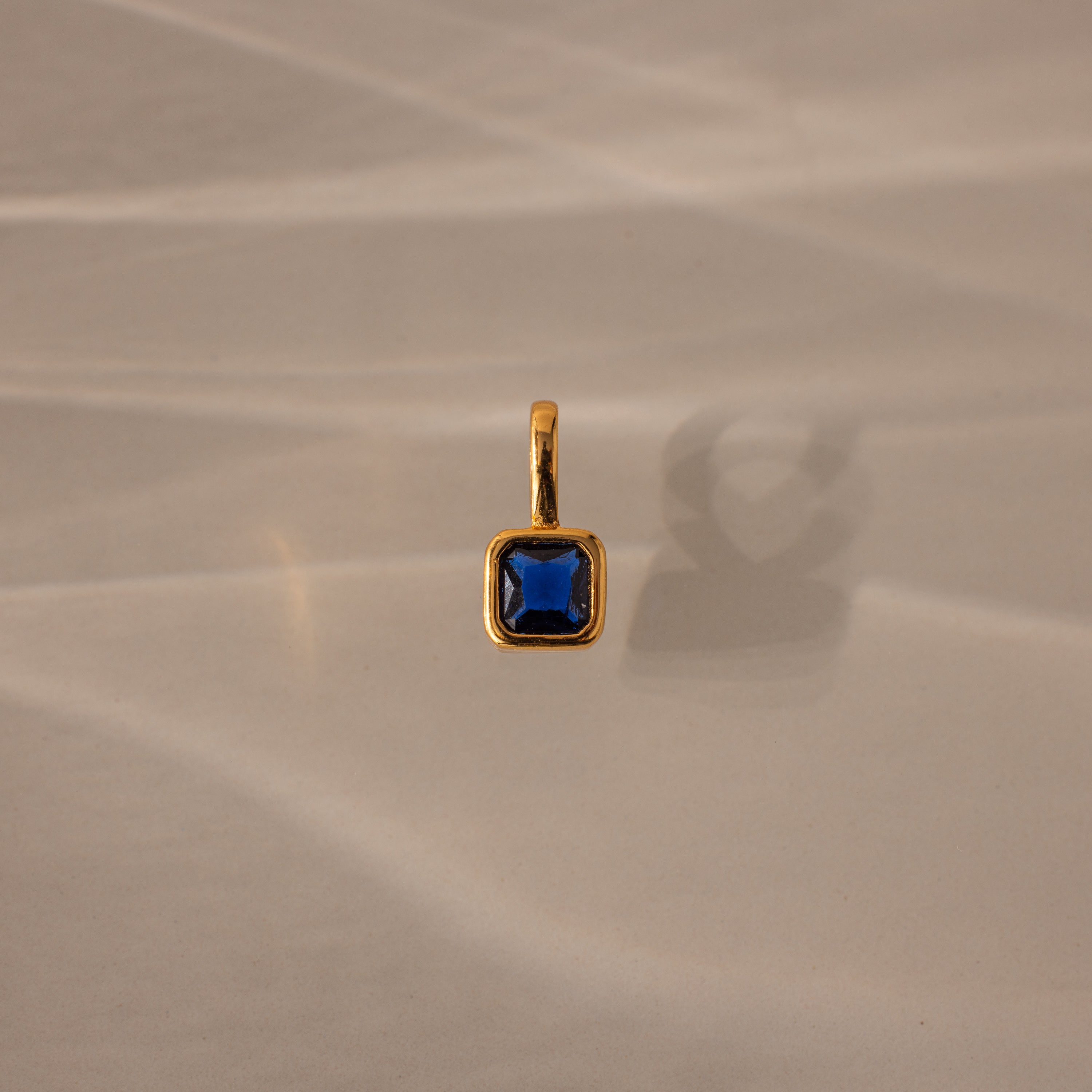 The Square Bezel Sapphire Charm, featuring a blue square gemstone set in a delicate gold frame, is ideal as a birthstone pendant and is shown resting on a beige surface with soft shadows.
