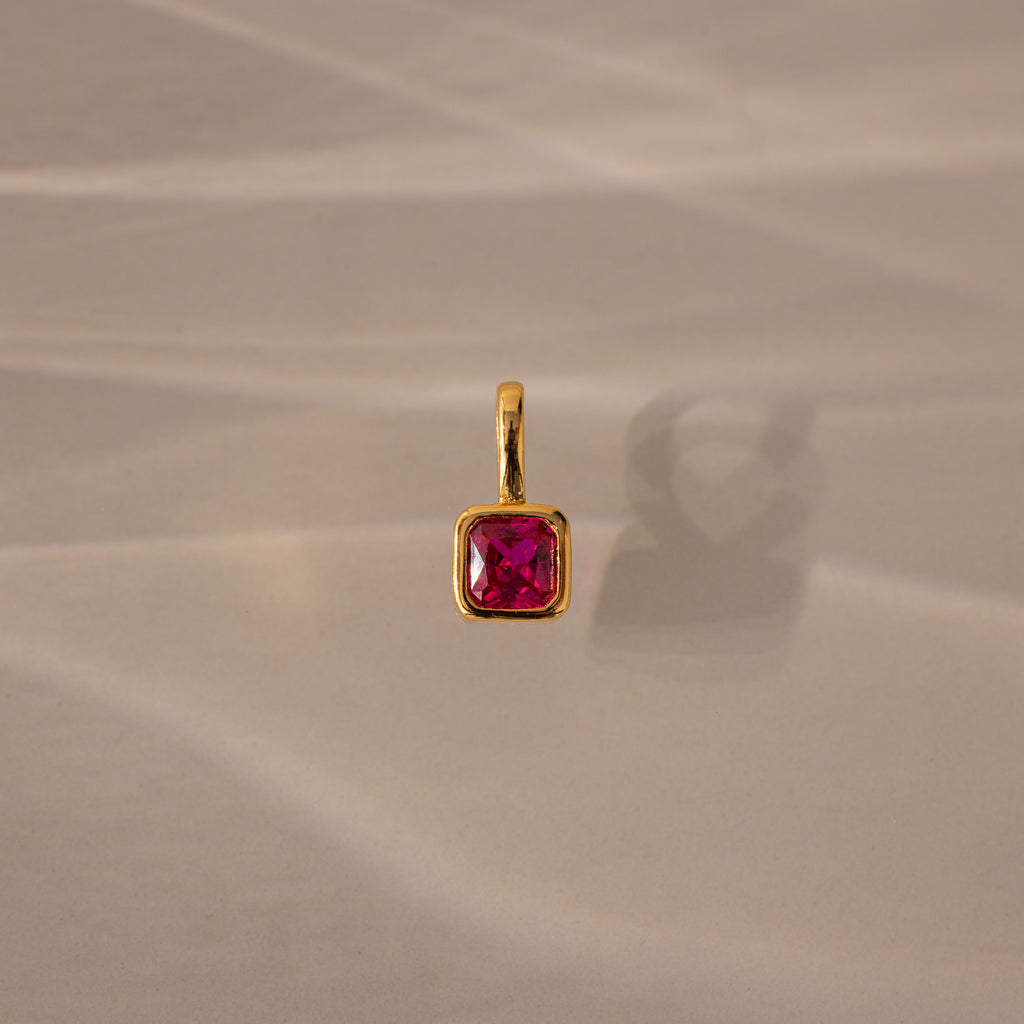 The Square Bezel Ruby Charm features a gold pendant with a square bezel and vibrant ruby, perfect as a birthstone charm for elegant jewelry lovers. Shown on a beige surface with soft shadows, it's ideal for any refined collection.