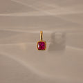 The Square Bezel Ruby Charm features a gold pendant with a square bezel and vibrant ruby, perfect as a birthstone charm for elegant jewelry lovers. Shown on a beige surface with soft shadows, it's ideal for any refined collection.