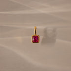The Square Bezel Ruby Charm features a gold pendant with a square bezel and vibrant ruby, perfect as a birthstone charm for elegant jewelry lovers. Shown on a beige surface with soft shadows, it's ideal for any refined collection.