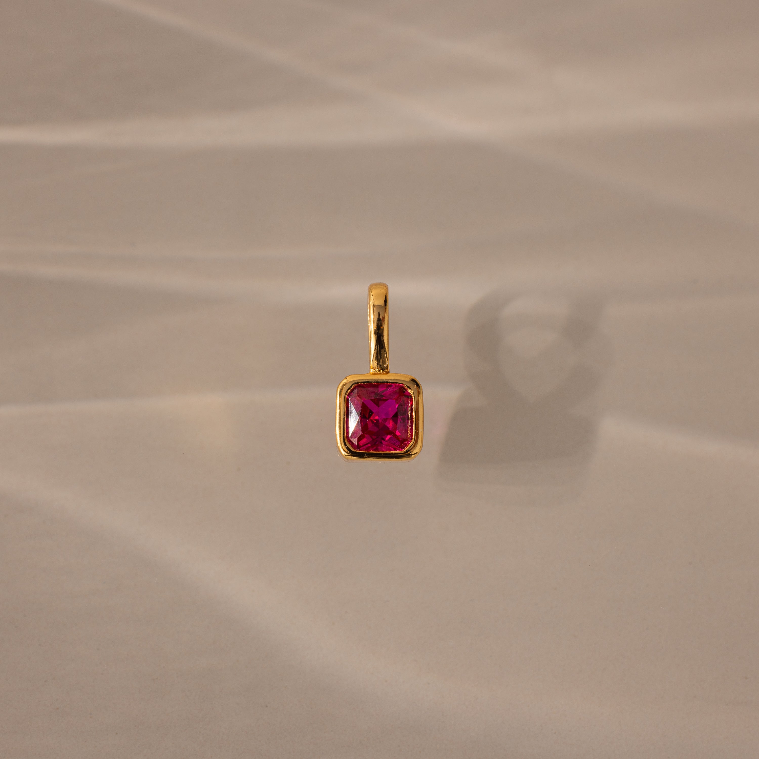 The Square Bezel Ruby Charm features a gold pendant with a square bezel and vibrant ruby, perfect as a birthstone charm for elegant jewelry lovers. Shown on a beige surface with soft shadows, it's ideal for any refined collection.
