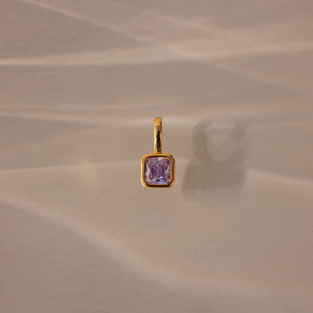 The Square Bezel Alexandrite Charm, featuring a square purple gemstone in a delicate gold frame, is displayed on a softly textured beige background.
