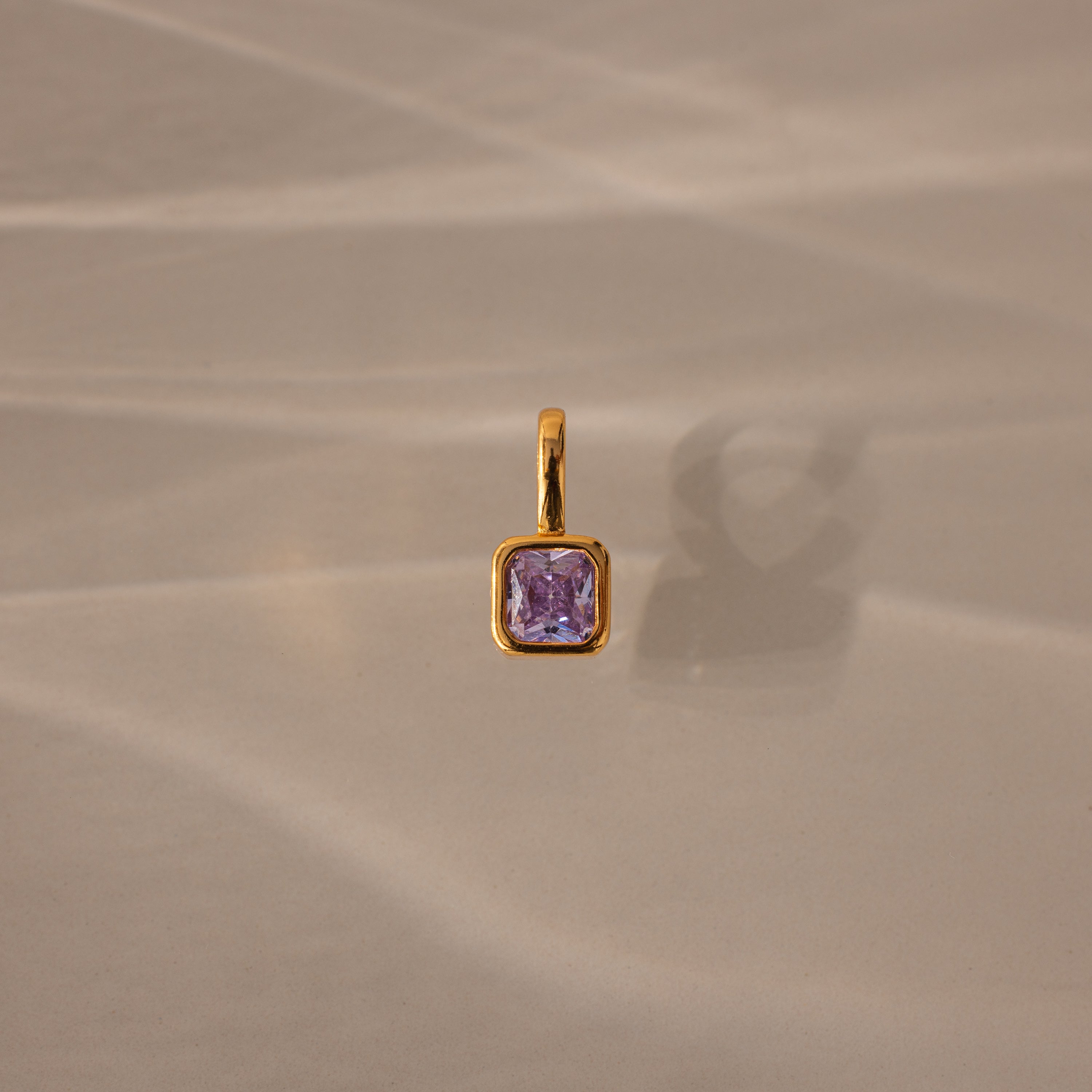 The Square Bezel Alexandrite Charm, featuring a square purple gemstone in a delicate gold frame, is displayed on a softly textured beige background.