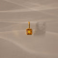 The Square Bezel Topaz Charm features a square yellow gemstone in a gold frame, casting a soft shadow on a beige surface for an elegant jewelry accent.