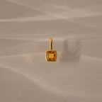 The Square Bezel Topaz Charm features a square yellow gemstone in a gold frame, casting a soft shadow on a beige surface for an elegant jewelry accent.