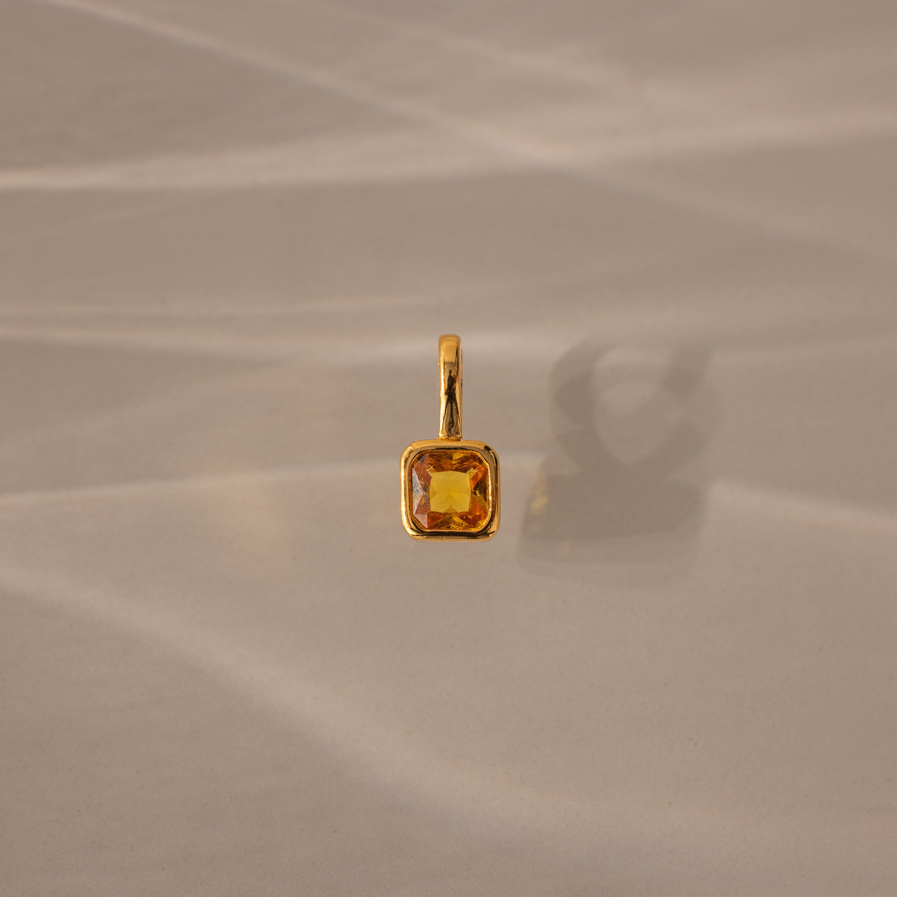 The Square Bezel Topaz Charm features a square yellow gemstone in a gold frame, casting a soft shadow on a beige surface for an elegant jewelry accent.