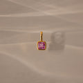 The Square Bezel Pink Tourmaline Charm features a pink tourmaline gemstone in a delicate gold frame, resting on a softly lit beige surface—an elegant and refined jewelry piece.