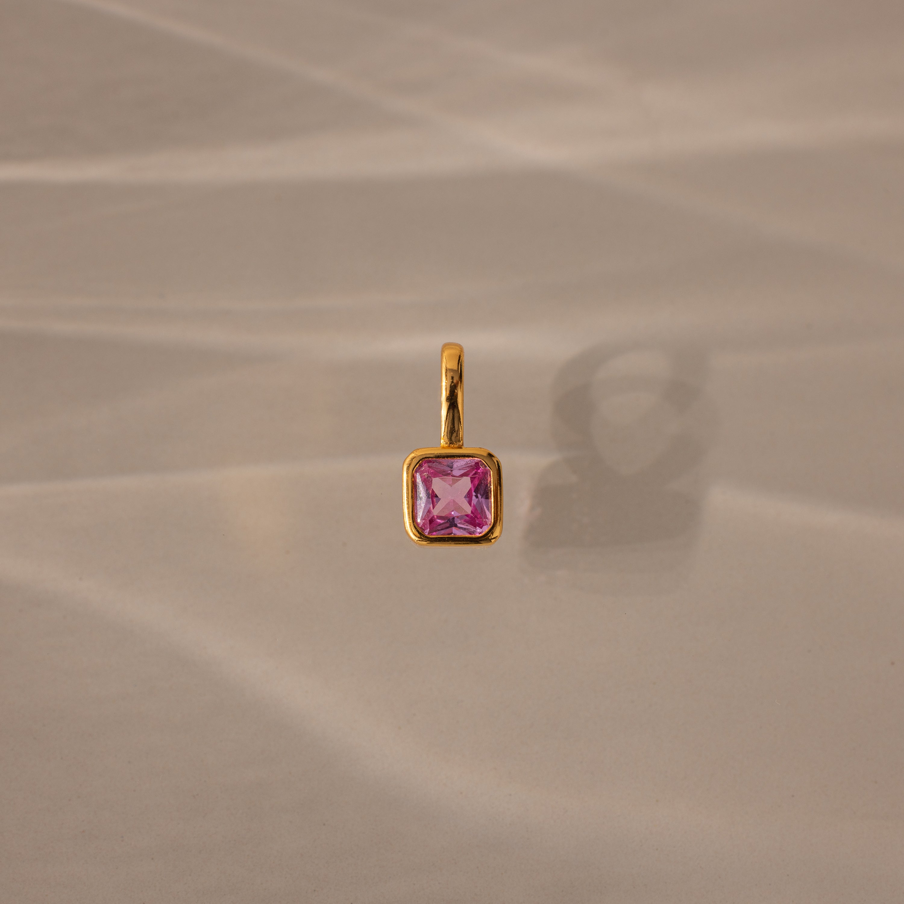 The Square Bezel Pink Tourmaline Charm features a pink tourmaline gemstone in a delicate gold frame, resting on a softly lit beige surface—an elegant and refined jewelry piece.