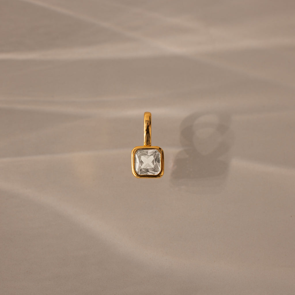 The Square Bezel Diamond Charm, a gold pendant featuring a square diamond, rests on a beige, softly textured background—an elegant piece for any gemstone jewelry collection.