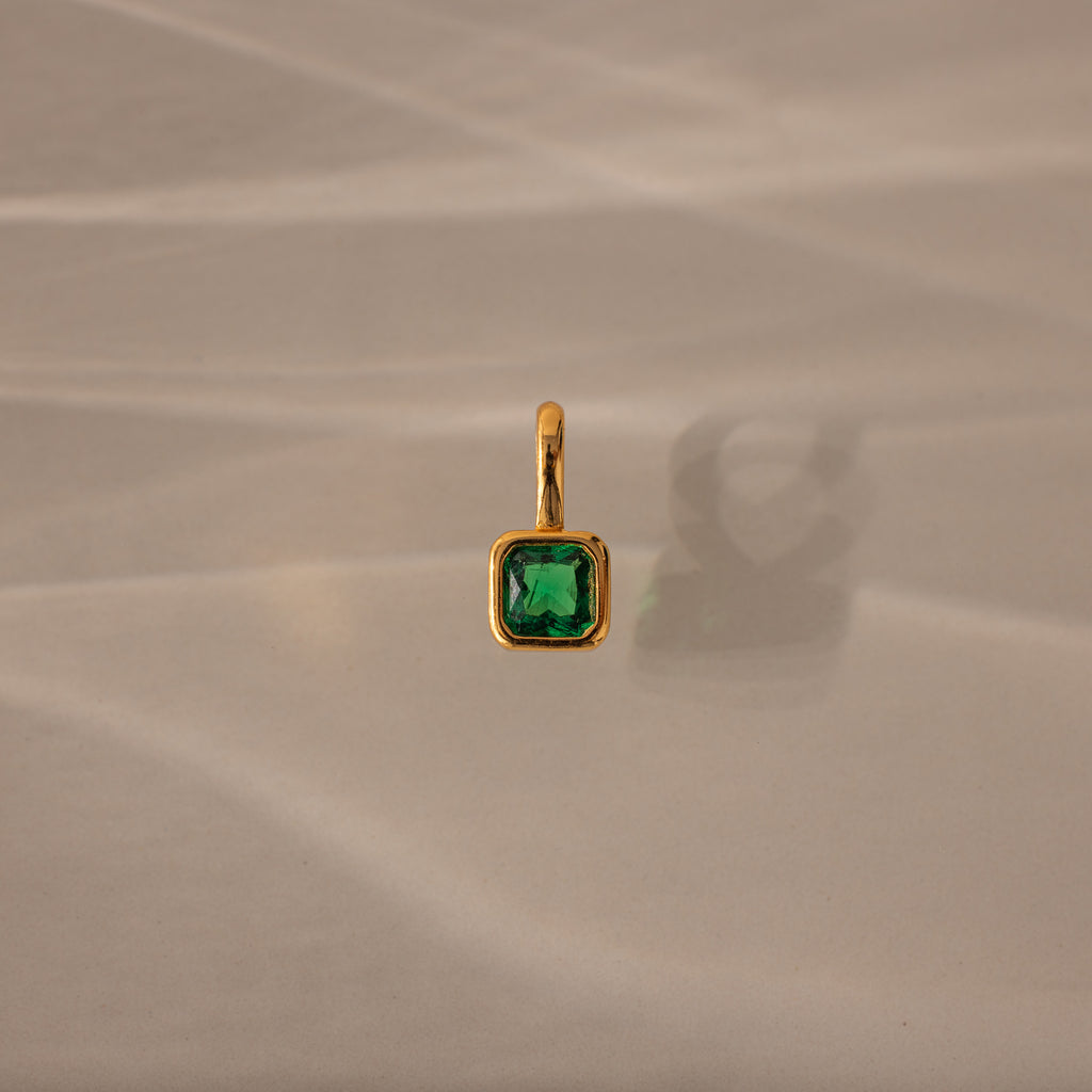 Square Bezel Emerald Charm: a gold pendant featuring a square green gemstone set in a gold frame, displayed on a beige textured background with a soft shadow.