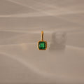 Square Bezel Emerald Charm: a gold pendant featuring a square green gemstone set in a gold frame, displayed on a beige textured background with a soft shadow.