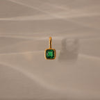 Square Bezel Emerald Charm: a gold pendant featuring a square green gemstone set in a gold frame, displayed on a beige textured background with a soft shadow.
