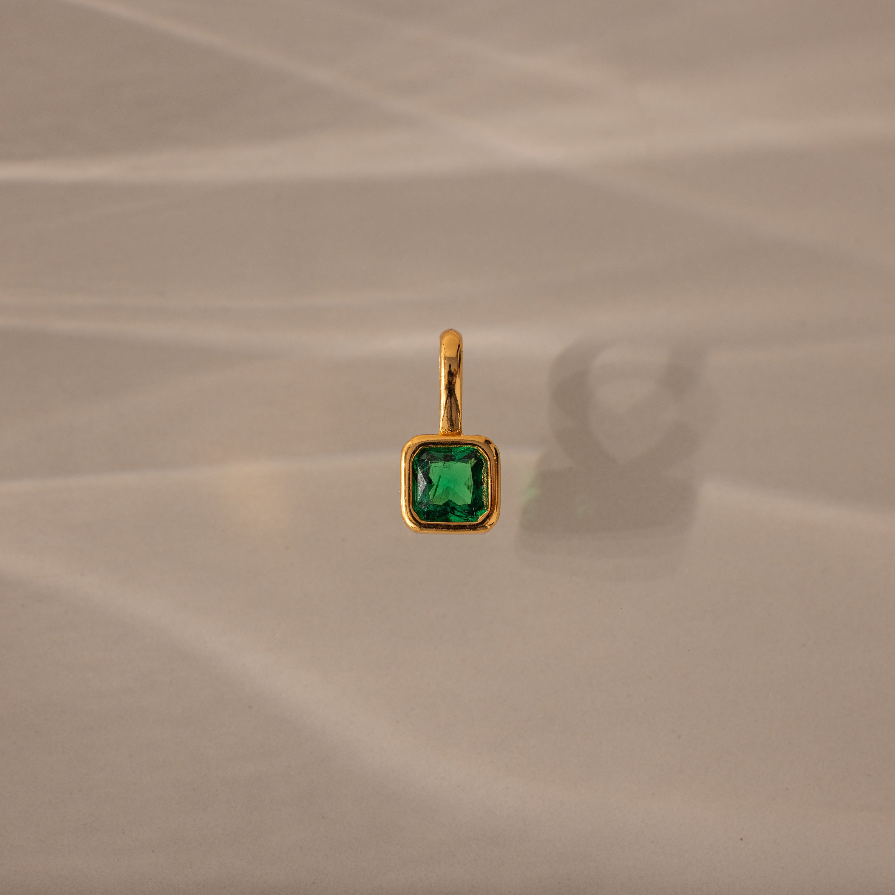 Square Bezel Emerald Charm: a gold pendant featuring a square green gemstone set in a gold frame, displayed on a beige textured background with a soft shadow.