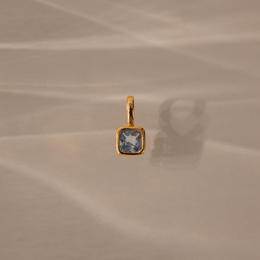 The Square Bezel Aquamarine Charm, a gold pendant featuring a faceted square blue gemstone, is displayed on a beige textured surface.
