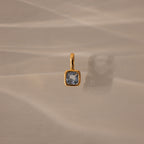 The Square Bezel Aquamarine Charm, a gold pendant featuring a faceted square blue gemstone, is displayed on a beige textured surface.