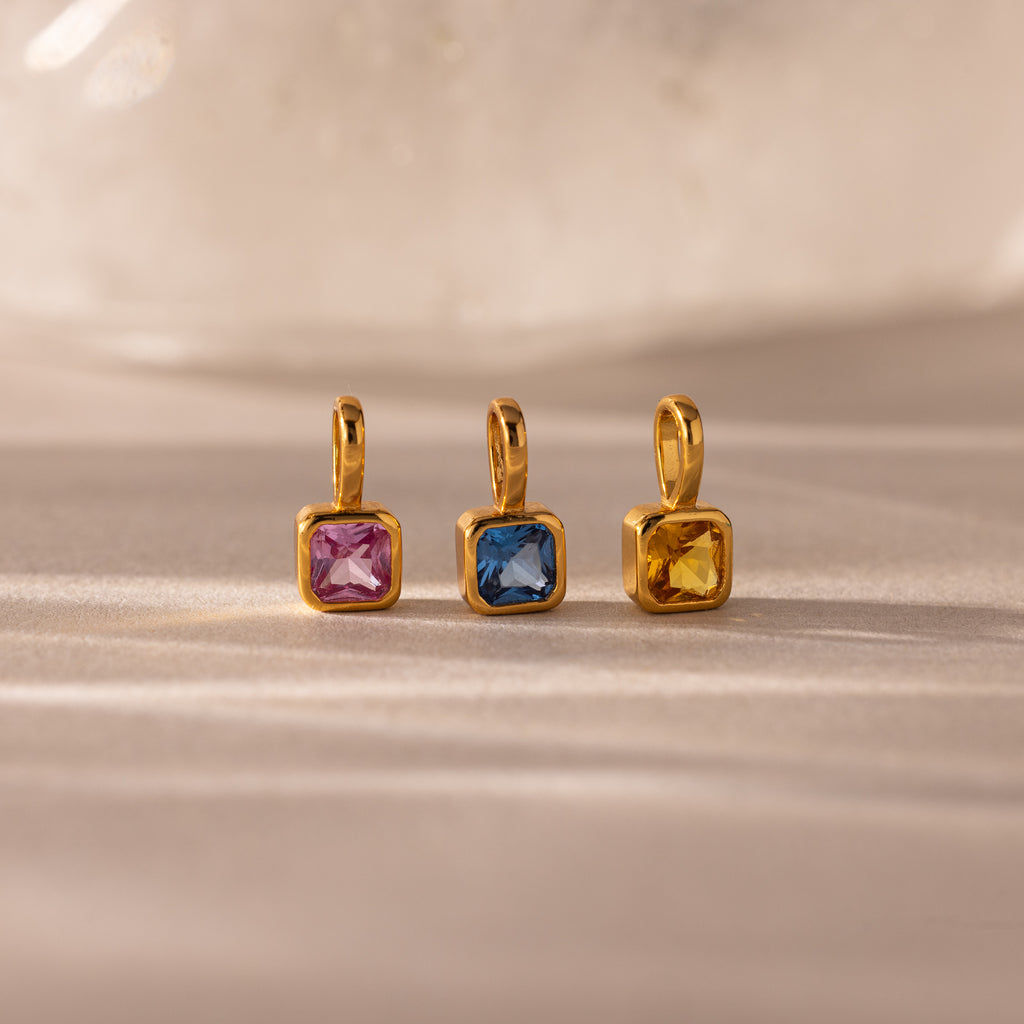 Three Square Bezel Birthstone Charms, each featuring a gold frame with square-cut pink, blue, and yellow gemstones, are displayed on a neutral surface—an elegant showcase of birthstone jewelry.
