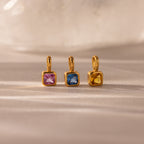 Three Square Bezel Birthstone Charms, each featuring a gold frame with square-cut pink, blue, and yellow gemstones, are displayed on a neutral surface—an elegant showcase of birthstone jewelry.