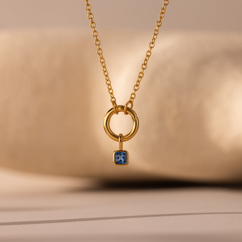 The Square Bezel Blue Zircon Charm is an elegant gold necklace featuring a circular pendant with a small square blue zircon gemstone set in a delicate gold frame, beautifully displayed against a soft background.