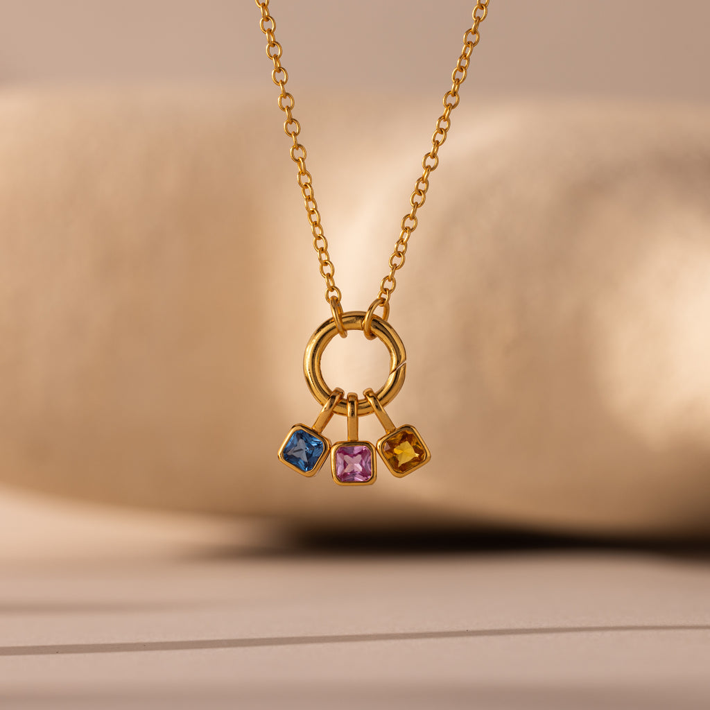 The Square Bezel Sapphire Charm features three square gemstones—blue, pink, and yellow—set in gold on a circular pendant. This elegant gold necklace is a stunning piece of gemstone jewelry, perfect for any occasion.