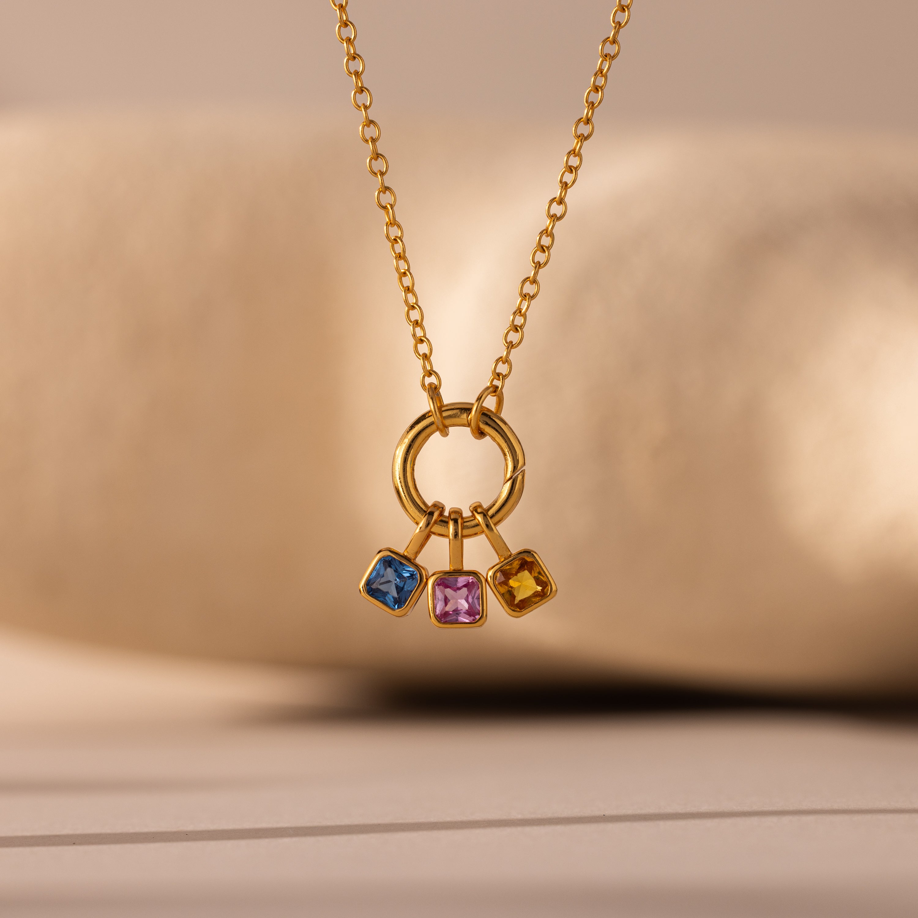 The Square Bezel Peridot Charm is a gold necklace featuring a circular gold frame pendant with three square gemstones—blue, pink, and yellow—for a bold gemstone jewelry statement.