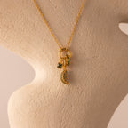 The Square Bezel Peridot Charm features three green peridot gemstones set in gold frames, displayed on a textured beige bust—an elegant and timeless piece of gemstone jewelry.