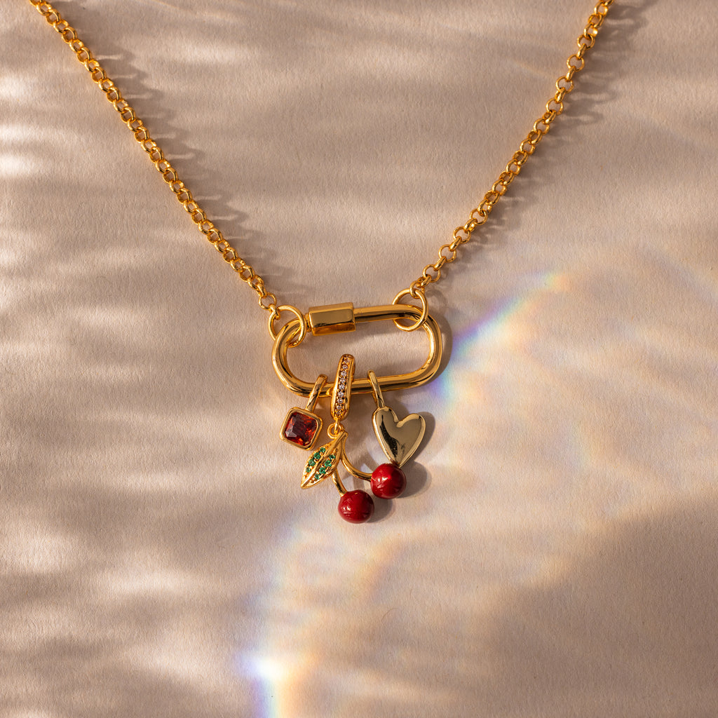 Gold necklace featuring the Square Bezel Ruby Charm, along with leaf, cherry, and heart charms, displayed on a textured surface with light reflections.