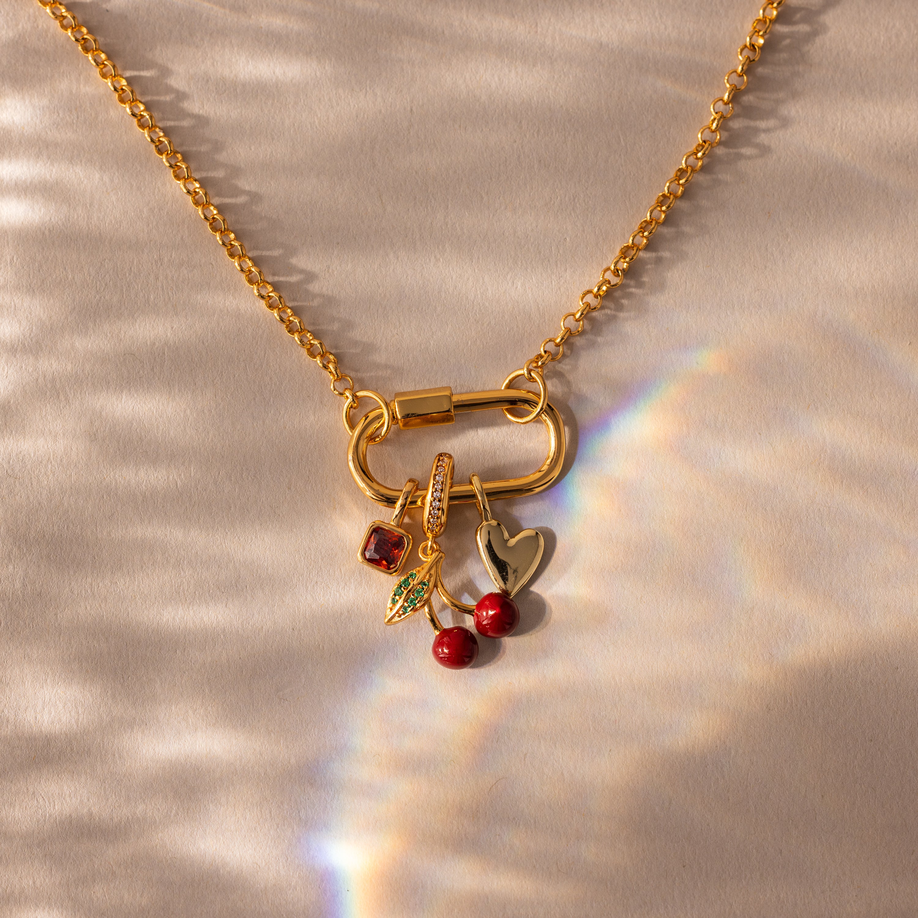 Gold necklace featuring the Square Bezel Ruby Charm, along with leaf, cherry, and heart charms, displayed on a textured surface with light reflections.