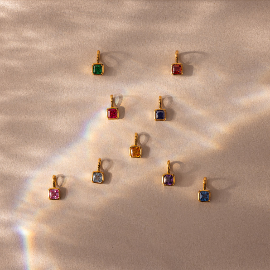 Nine Square Bezel Emerald Charms with gold frames are arranged on a beige, sunlit background, each one sparkling like a unique gemstone pendant.