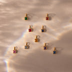 The Square Bezel Aquamarine Charm features a faceted aquamarine gemstone in a gold frame, displayed on a textured beige surface and shimmering in rainbow light.