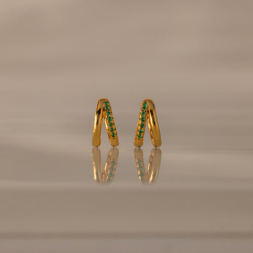 The Amelia Duo Emerald Huggies are modern gold hoop earrings with vibrant green gemstones, standing upright with their reflection shimmering on a smooth, neutral surface.