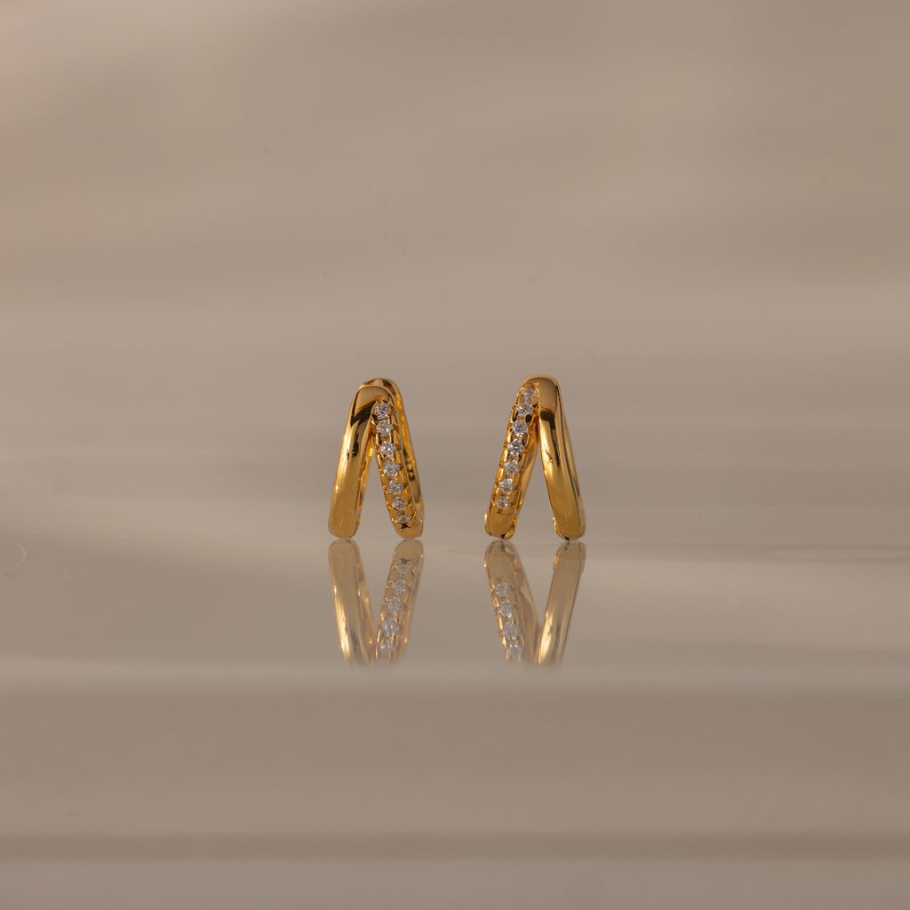 Amelia Duo Diamond Huggies – gold V-shaped modern earrings featuring sparkling diamonds, showcased on a reflective beige surface.
