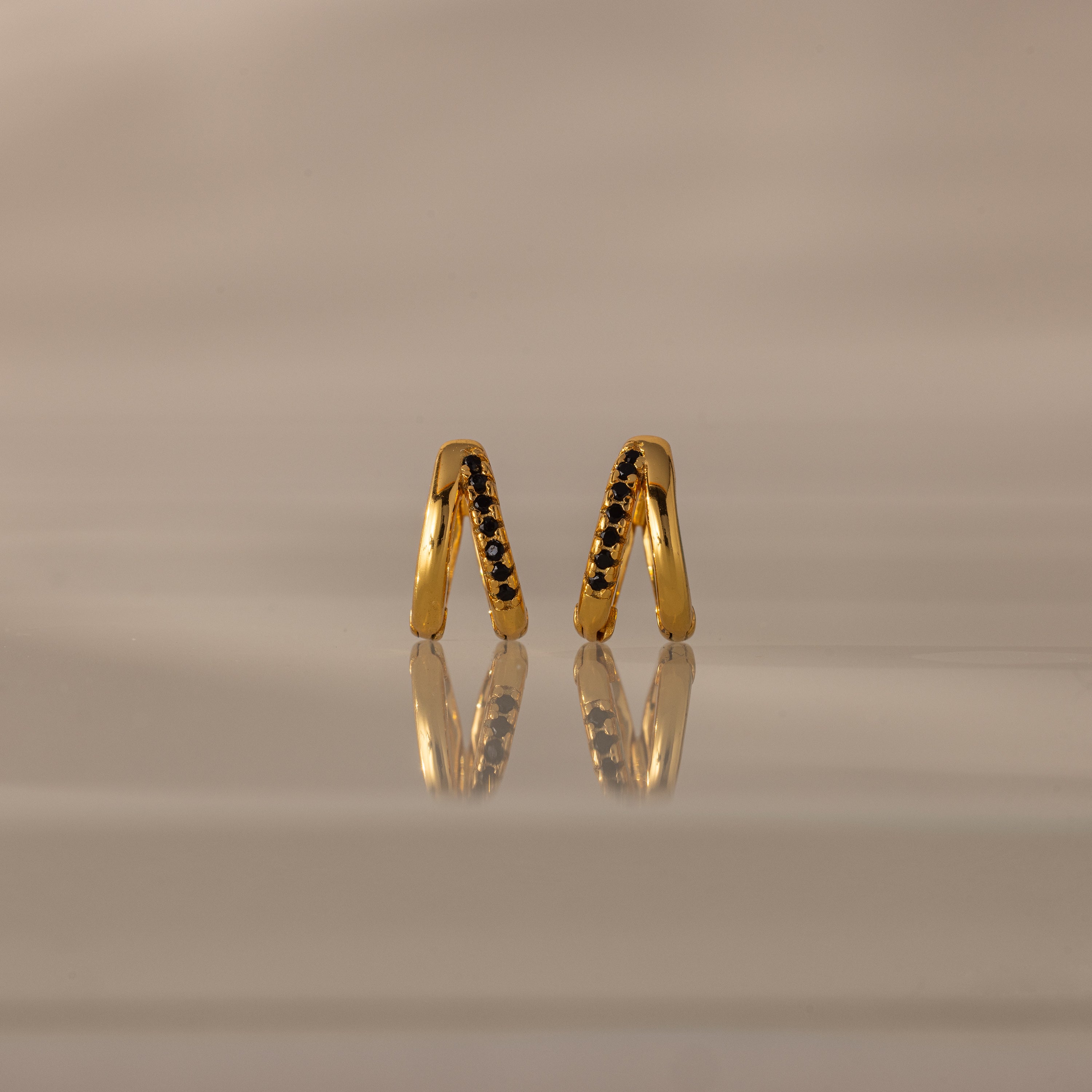 The Amelia Duo Onyx Huggies are modern V-shaped earrings featuring black onyx stones, elegantly reflected on a smooth, beige surface.