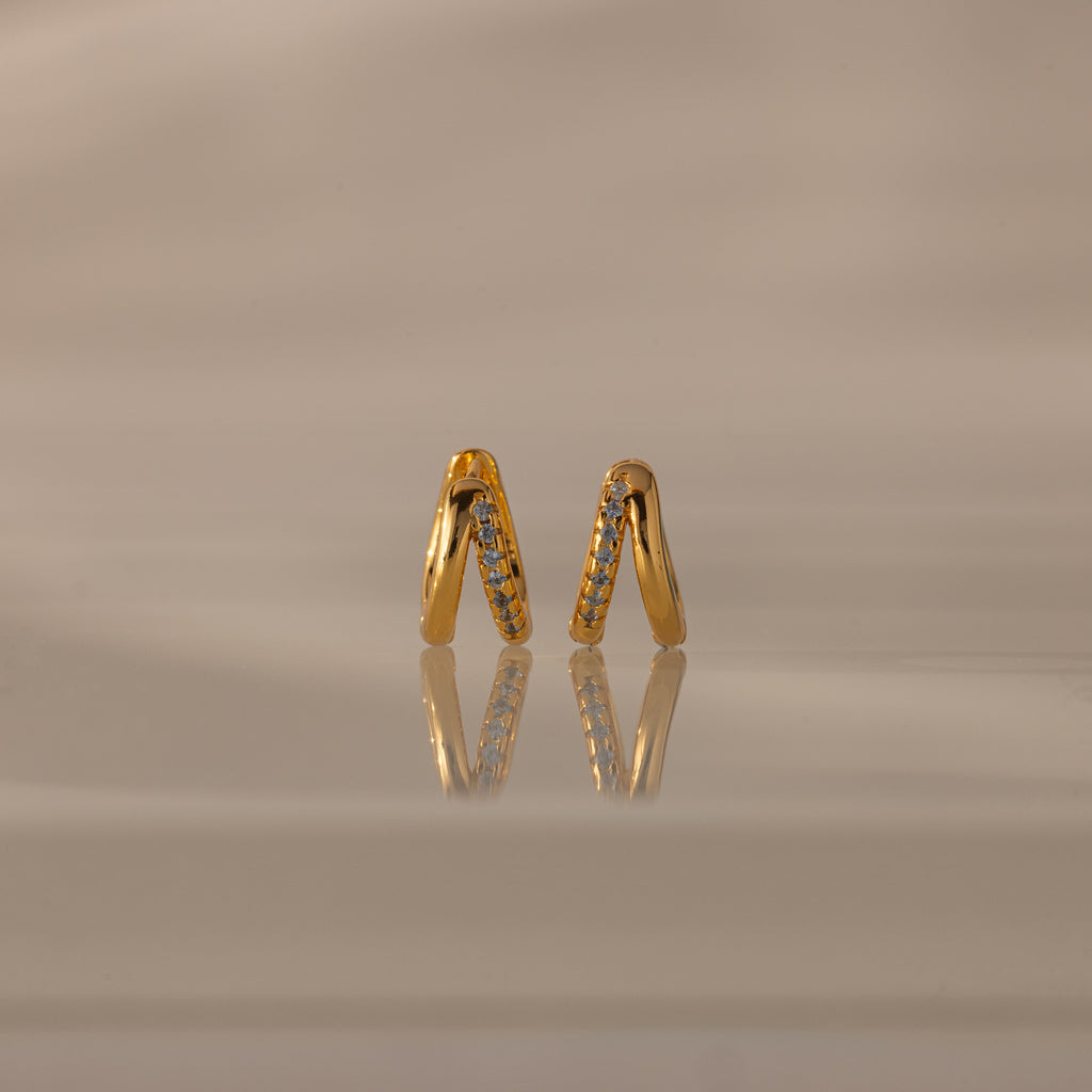 Amelia Duo Birthstone Huggies in Aquamarine (March), gold V-shaped modern earrings with small gemstones, displayed on a reflective beige surface.