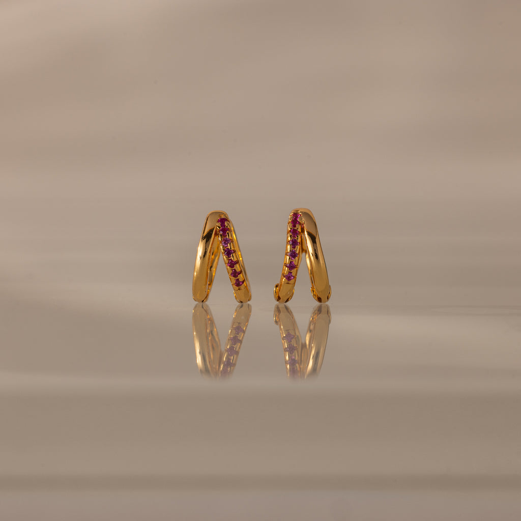 Amelia Duo Ruby Huggies—gold modern earrings with pink gemstones and intertwined hoops—stand upright on a reflective beige surface.
