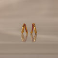 Amelia Duo Ruby Huggies—gold modern earrings with pink gemstones and intertwined hoops—stand upright on a reflective beige surface.